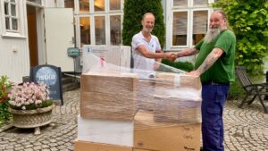 To menn håndhilser foran stabler med esker merket “Omega-3 kapsler” utenfor inngangen til isblå.no, klare for levering til Matsentralen.