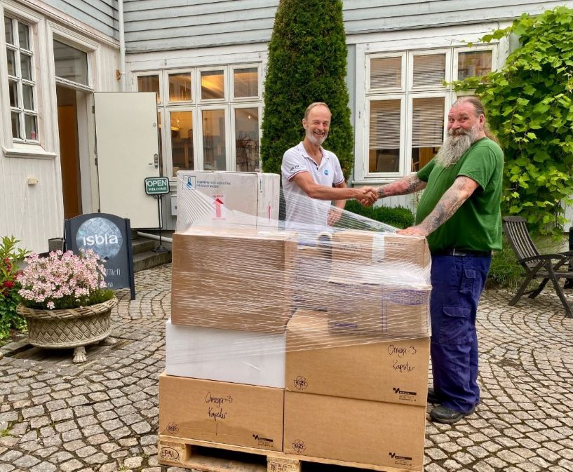 To menn håndhilser foran stabler med esker merket “Omega-3 kapsler” utenfor inngangen til isblå.no, klare for levering til Matsentralen.